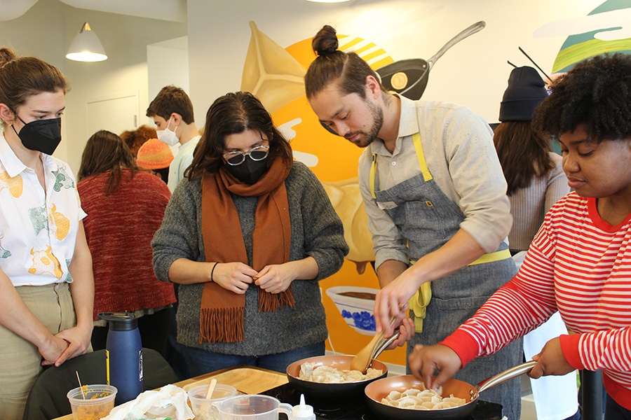 Several people stand around pans, cooking dumplings. One person wears a chef's apron.
