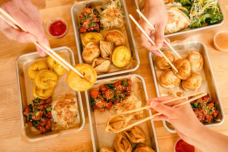 Overhead view of several small metal trays full of a variety of fried dumplings, with three hands each holding a dumpling in chopsticks above the trays.