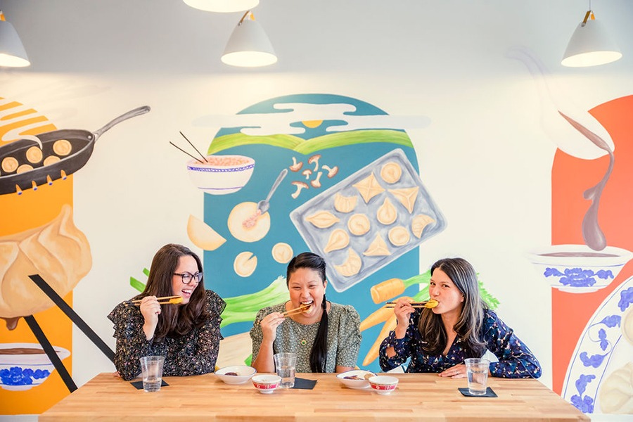 Three women sit at a table, smiling at eating dumplings with chopsticks in front of a mural depicting dumplings.
