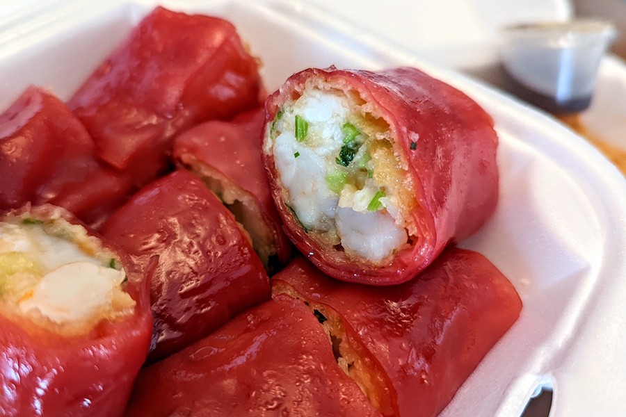 Closeup on a dim sum dish of bite-sized pieces of red noodles wrapped around crispy shrimp, served in a white styrofoam to-go container.