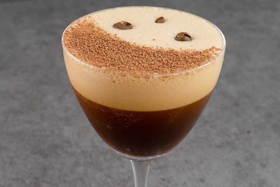 Closeup on a brown cocktail with a thick foamy top, garnished with a light brown powder and coffee beans.