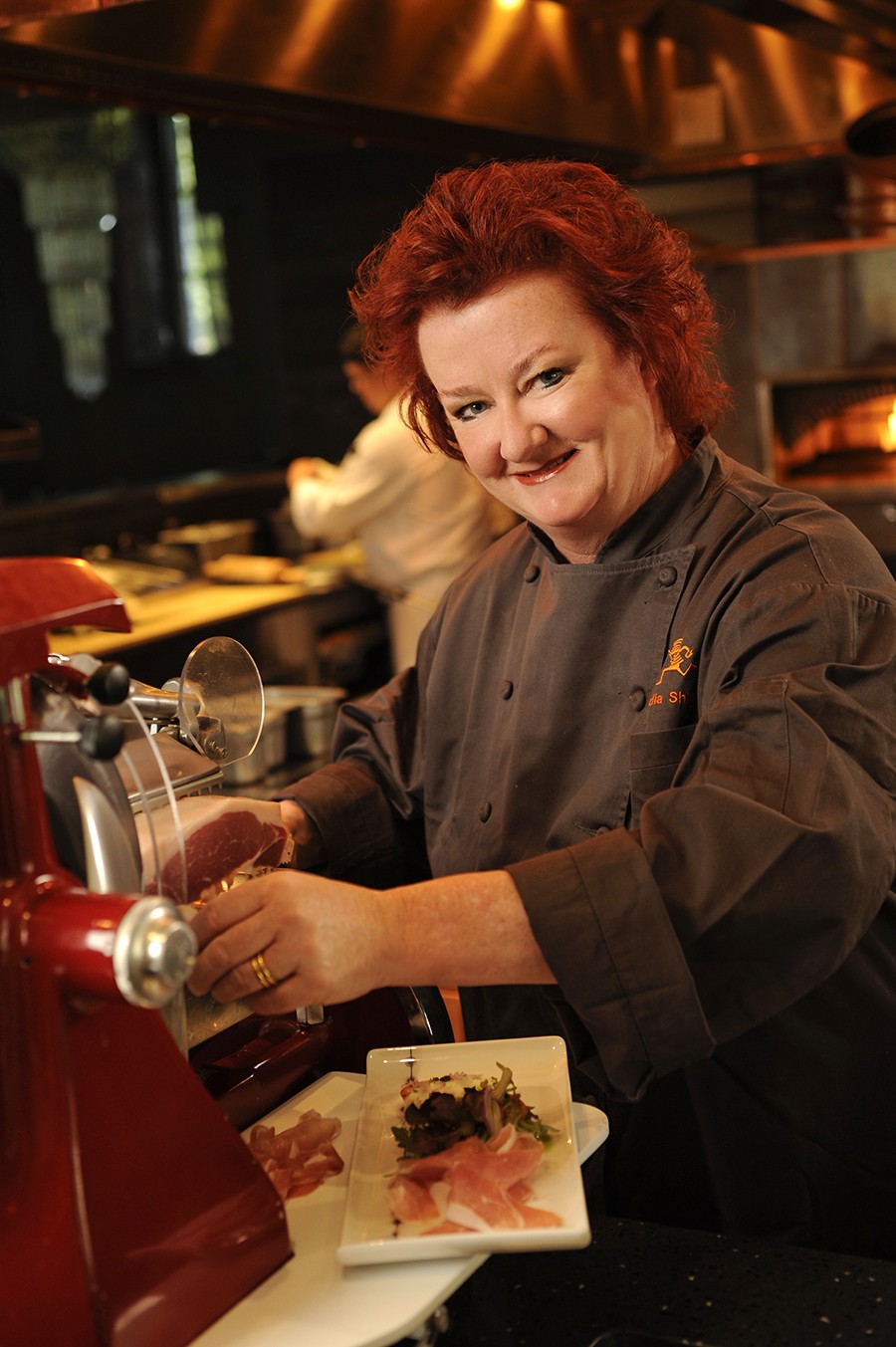A red-headed woman in chef clothes stands in a restaurant kitchen and smiles at the camera.