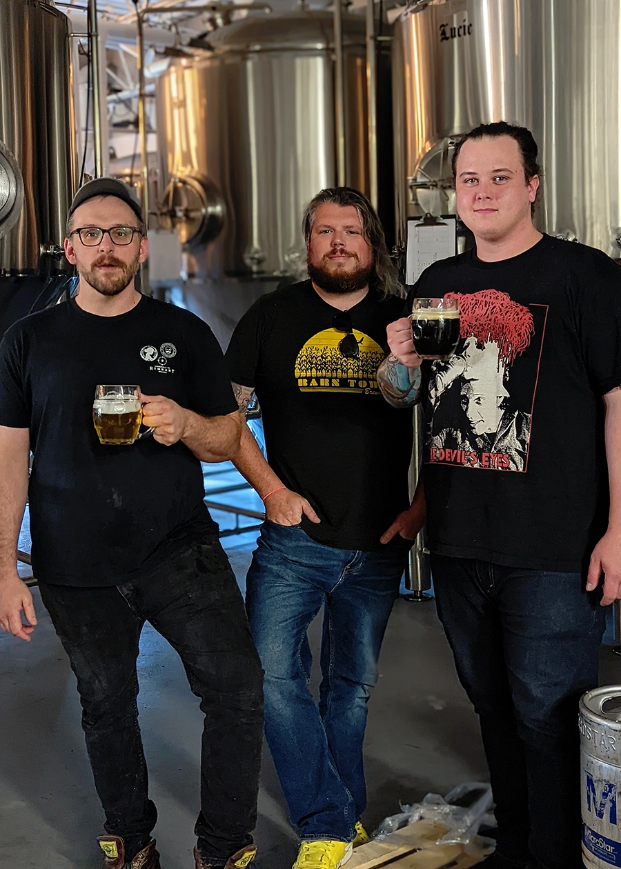Three men in jeans and black graphic t-shirts stand in front of brewing equipment.