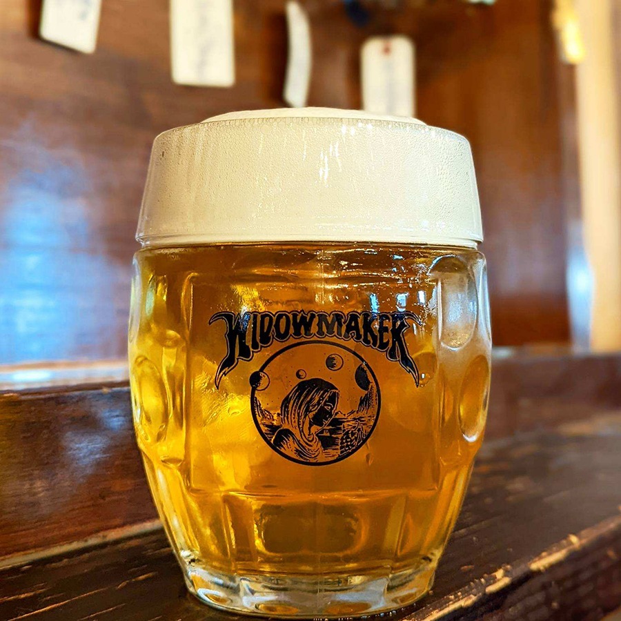 A glass of a golden beer has Widowmaker branding on it and sits on a wooden counter.