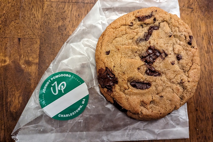 A chocolate chip cookie sits on a bag with a green sticker that says Johnny Pomodoro Charlestown, MA