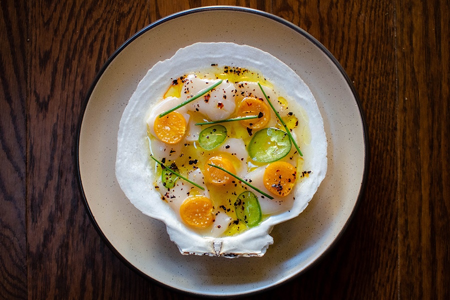 Overhead view of a scallop crudo, served in a scallop shell, garnished with bright yellow-orange gooseberries and green slices of jalapeno.