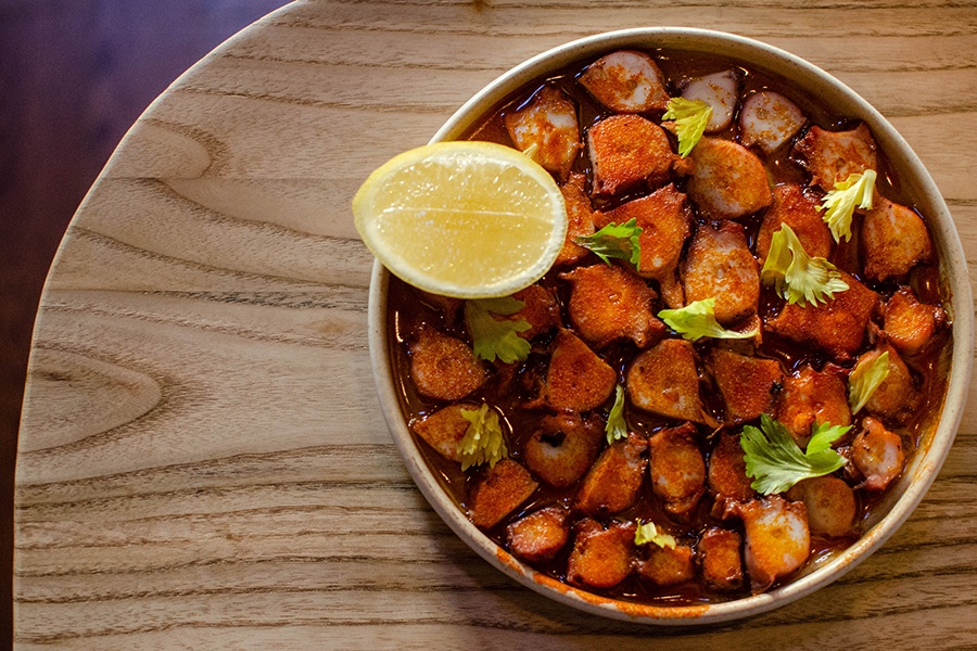 Overhead view of chopped octopus in a round plate, tossed in bright red seasoning and garnished with herbs and a lemon wedge.