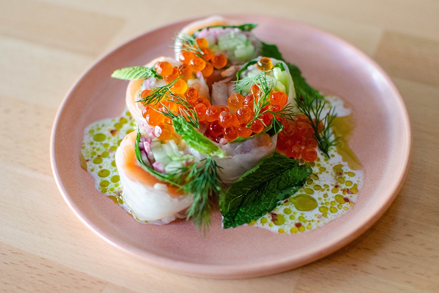 A fresh roll packed with cucumber and salmon is split into pieces and garnished with orange salmon roe and fresh herbs.
