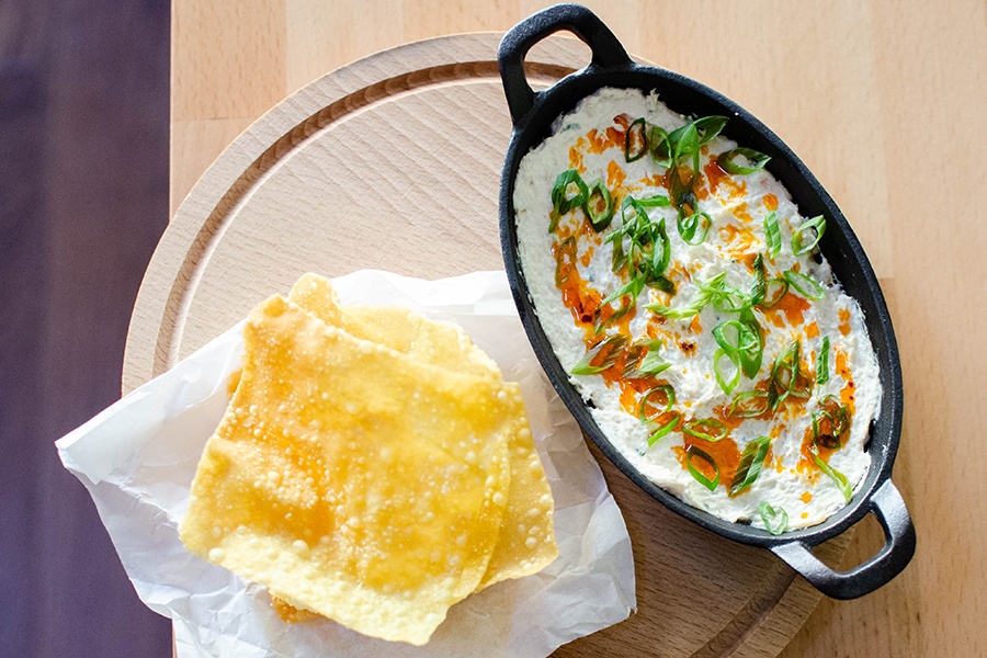 A cast-iron, oval-shaped pan is full of a thick white dip, garnished with scallions and an orange oil, with a side of crispy wontons.