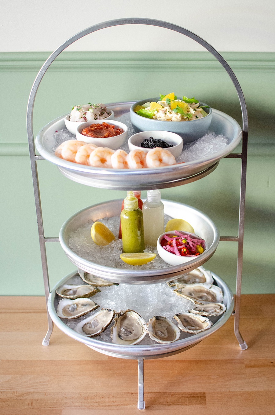 A three-story tower of silver trays holds raw bar items and sauces. It sits on a light wooden table in front of a pale green wall.