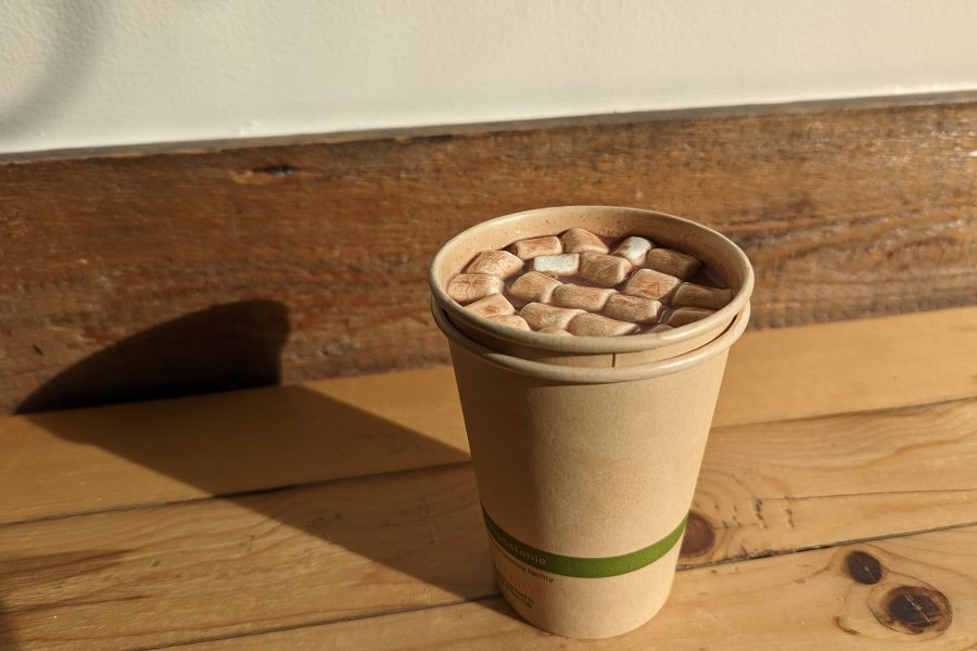 A paper cup is filled with hot chocolate and marshmallows and sits on a wooden counter.