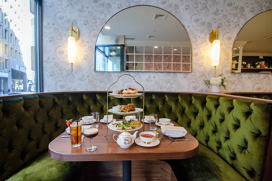 Afternoon tea service is set up at a wooden table in front of a green velvet banquet, with a half-circle mirror in the background.