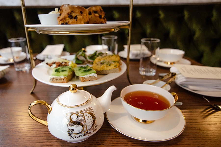 A teapot and delicate cup of tea sit on a table in front of tiers of savory afternoon tea bites and scones.