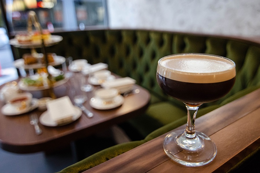 A delicate glass of a foam-covered espresso martini perches atop a classy green velvet restaurant banquet, with afternoon tea service visible in the background.