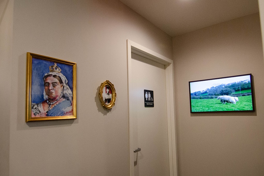 Photo of a restroom door in a restaurant. Near it, two pieces of artwork depict British queens, and a video screen shows sheep grazing in grass.