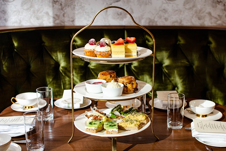 A three-tier setup of afternoon tea savory and sweet treats and scones sits on a wooden table in front of a green velvet banquette.