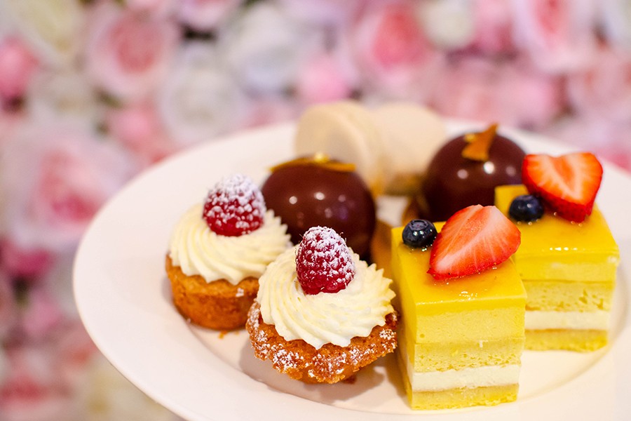 A plate of four types of afternoon tea pastries, including bright yellow layer-cake squares, sit on a plate in front of a background of pink and white roses.