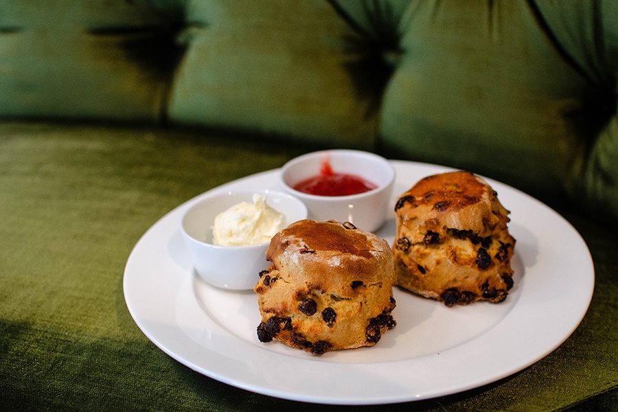 Two puffy British-style scones sit on a plate with two small bowls of condiments, a thick cream and a bright red jam.