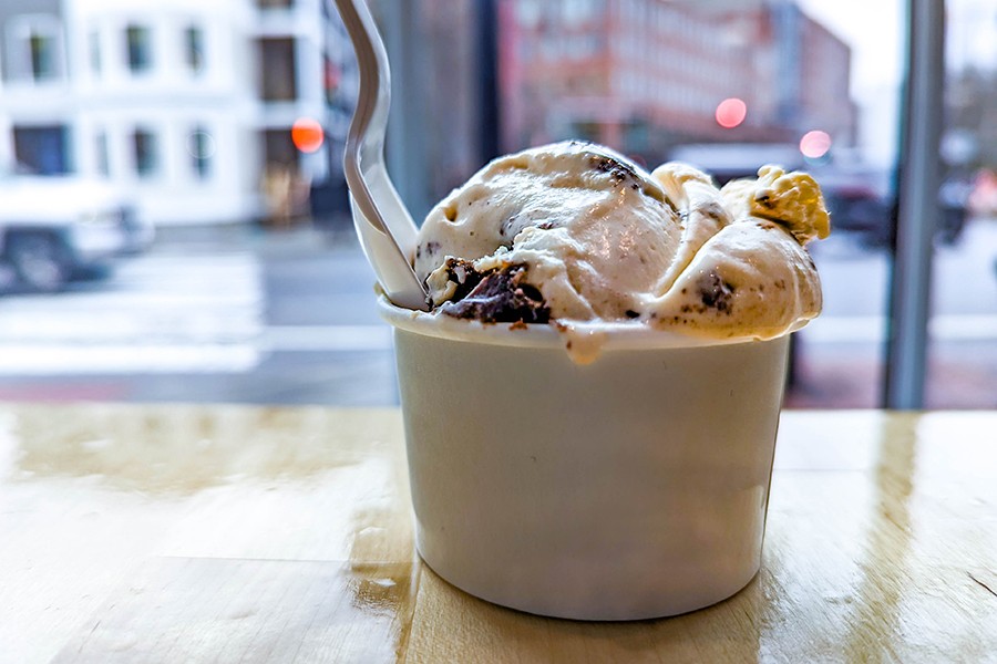 A scoop of an off-white ice cream with brownie chunks in a paper cup, with a white plastic spoon.