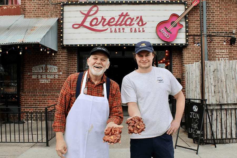 Two smiling men, one in a white apron, stand in front of a restaurant with signage reading Loretta's Last Call, each holding a big apple fritter.