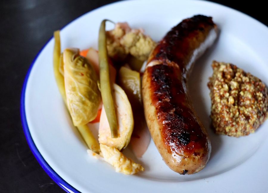 A single fat sausage sits on a white plate with a smear of whole-grain mustard and some pickled veggies.