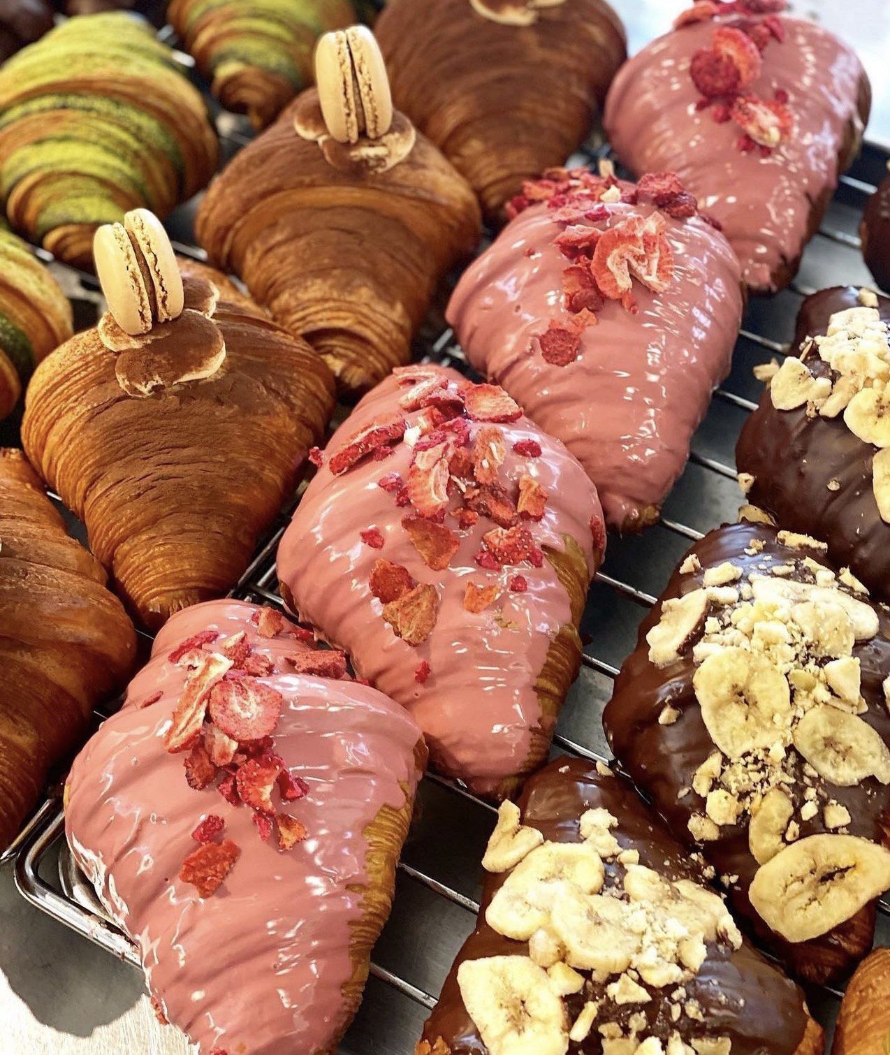 Rows of croissants are topped in a few different ways, with some completely covered in pink glaze, others in chocolate with banana chips, and more.