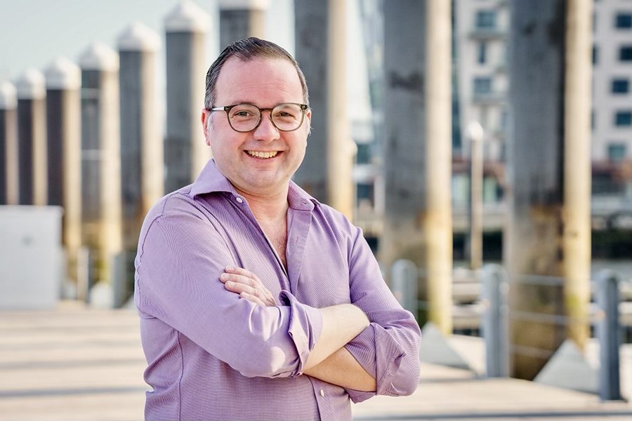 A man in glasses and a lavender button-down shirt stands, smiling and arms crossed, in the sun on a dock.
