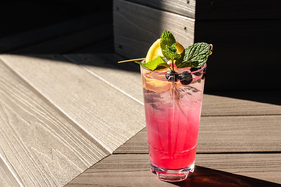A light pink cocktail in a pint glass is garnished with blueberries, mint, and a lemon slice.