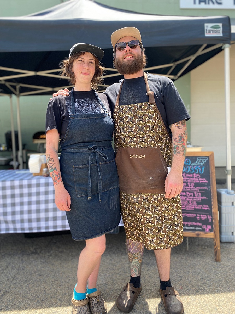 A man and a woman wearing chef's aprons and baseball caps smile, standing in front of an outdoor food pop-up setup.