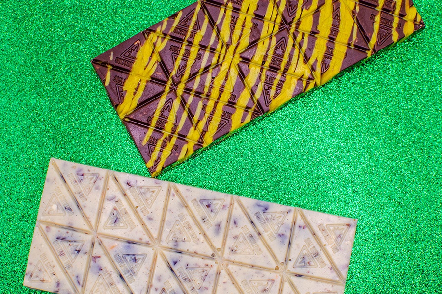 Two chocolate bars are on a sparkly green background. One is milk chocolate with vibrant yellow squiggles, and one is white chocolate with bits of cherries.