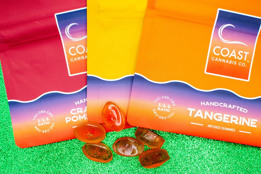Three bags of THC-infused gummies in bold red, orange, and yellow tones are displayed on a sparkly green background, with a handful of oval-shaped gummies visible.