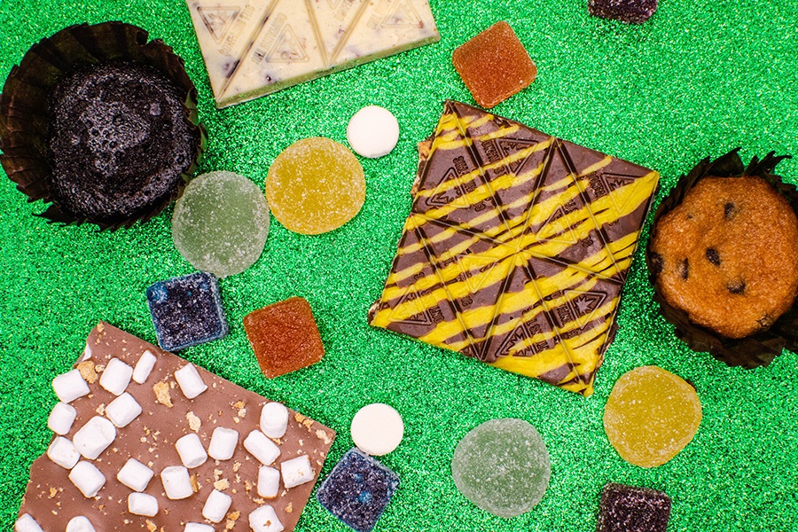 A spread of various THC-infused chocolates, gummies, mints, and baked goods on a sparkly green background.
