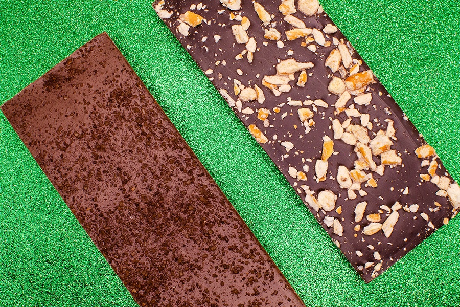 Two chocolate bars are displayed on a sparkly green background, one of which is covered with crushed pretzels.