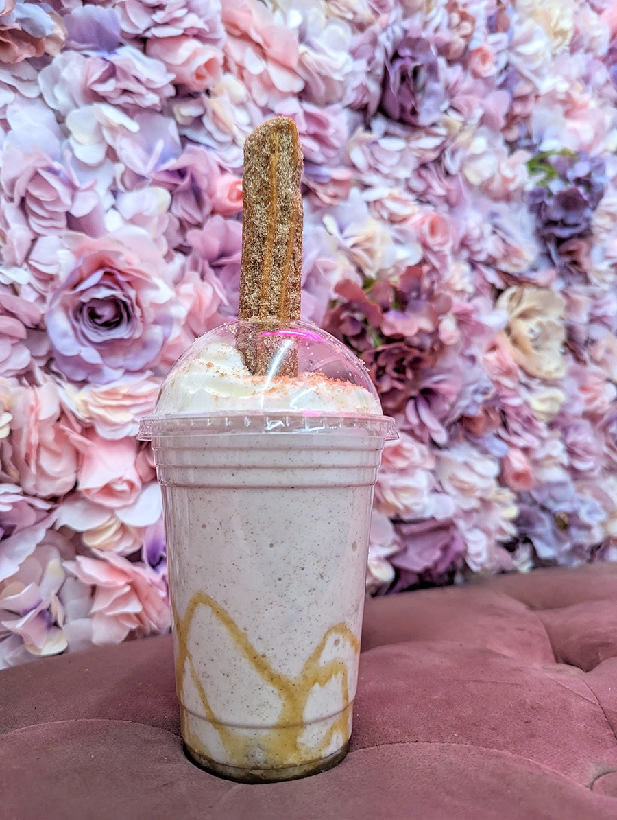 A milkshake is topped with whipped cream and a churro, photographed in front of a pink flower-covered wall.