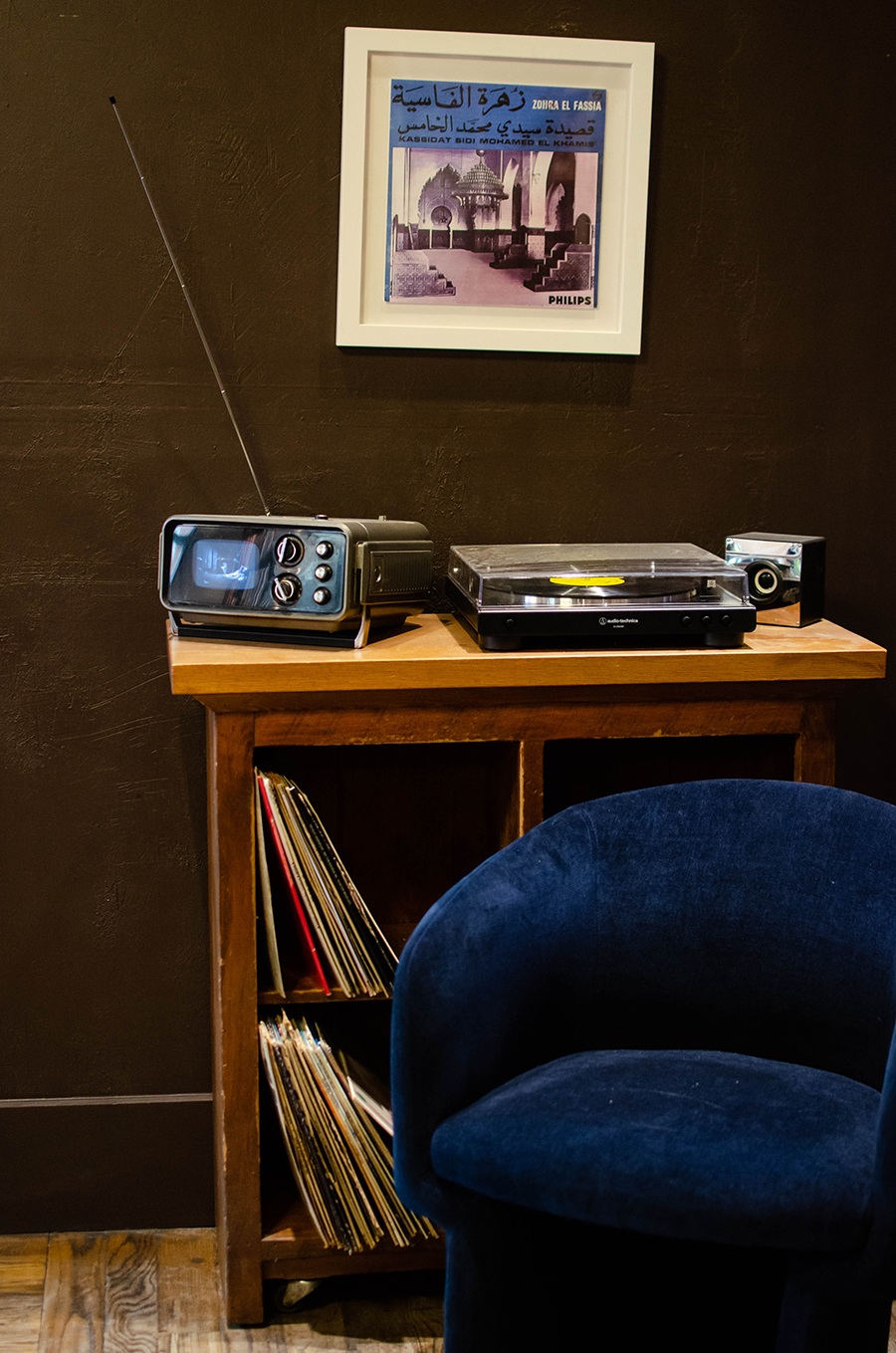 A blue velvet chair sits in front of a bookshelf with records, a record player, and a tiny old television.