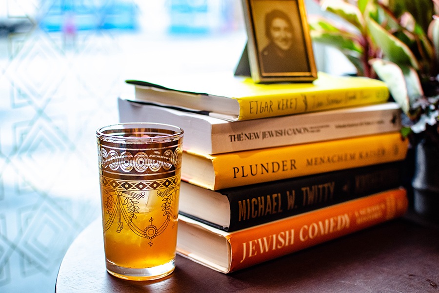 A brown cocktail in a delicate Moroccan tea glass with gold detailing sits next to a pile of books by Jewish authors.