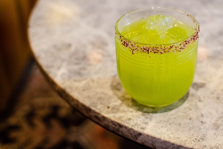 A greenish-yellow cocktail with a red salt rim sits on a marble table.