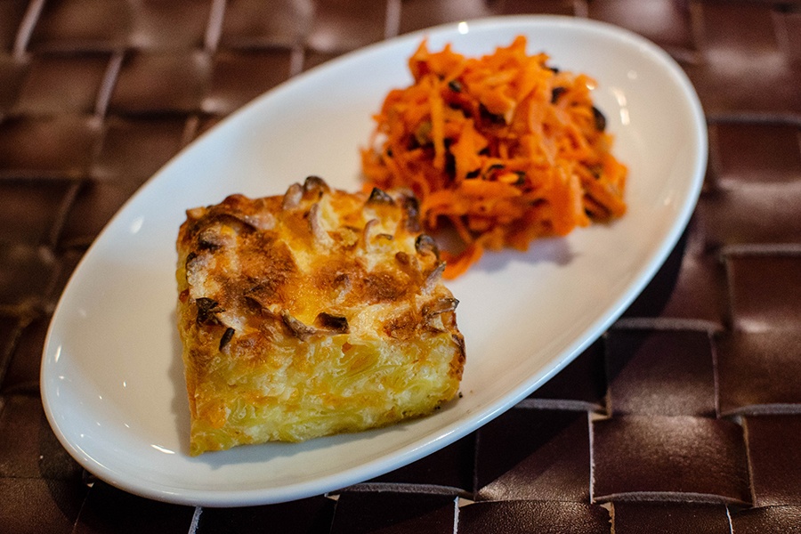 A square of golden-brown, cheesy noodle kugel is accompanied by a sliced carrot slaw.