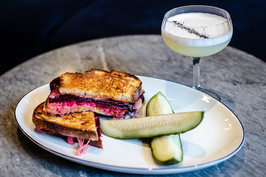 A sandwich with bright purple beets and a side of pickles is accompanied by a foamy cocktail with a stripe of poppyseeds as garnish.