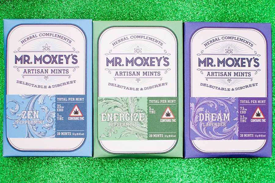 Three boxes of Mr. Moxey's Artisan Mints, each in a pastel blue, purple, or green, are lined up on a sparkly green background.