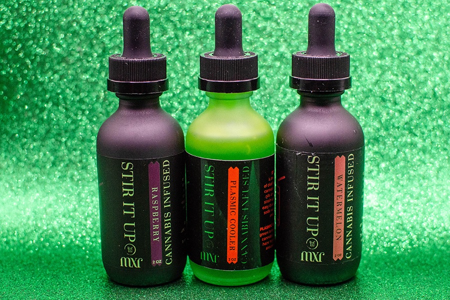 Three small bottles, two black and one bright green, are filled with a liquid and displayed on a sparkly green background.