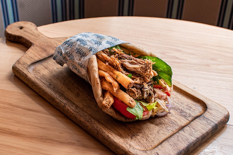 A fluffy pita is stuffed with pork, fries, tomato, and greens, presented on a wooden cutting board.