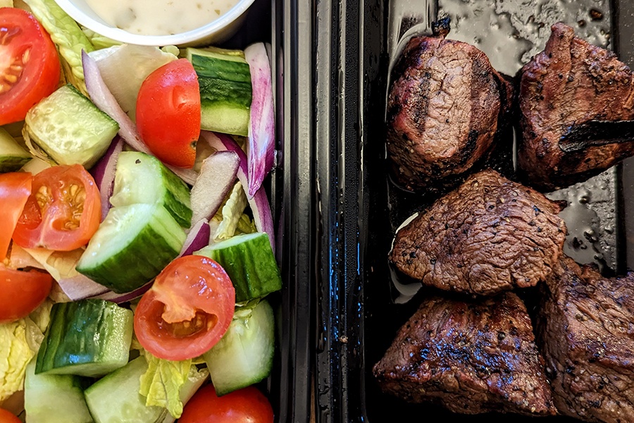 Overhead view of two takeout containers, one with salad and one with thick, grilled steak tips.