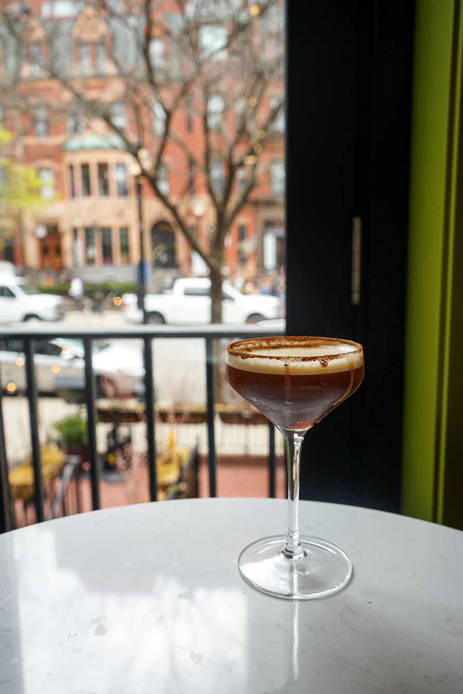 An espresso martini sits on a table in front of a large window looking out onto Boston's Newbury Street.