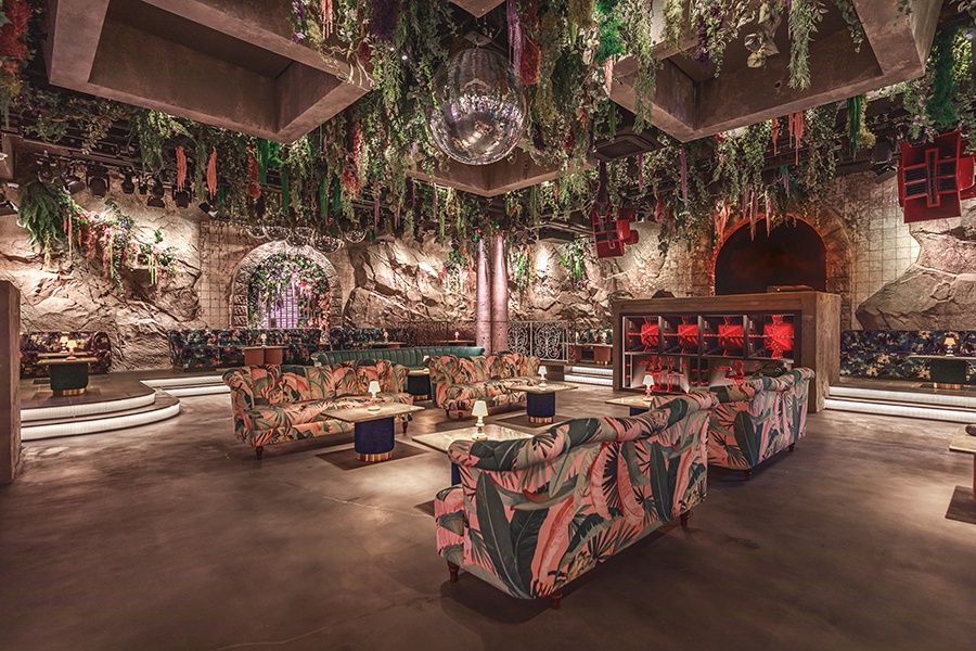 A nightclub looks like a cave with stone walls and draping plants. Lounge seating features tropical plant patterns.