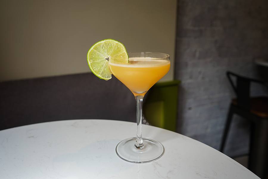 An orange cocktail served in a coupe glass is garnished with a lime slice.