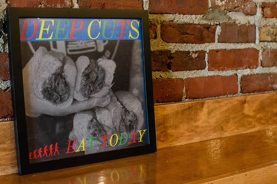 A piece of art that says Deep Cuts Eat Today, modeled after the Gorilla Biscuits Start Today album cover, is propped up on a wooden counter top against a brick wall.