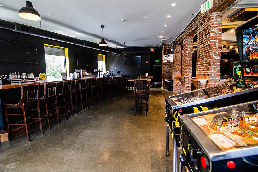 An empty bar features black walls with bright yellow accents, brick walls, and pinball machines.