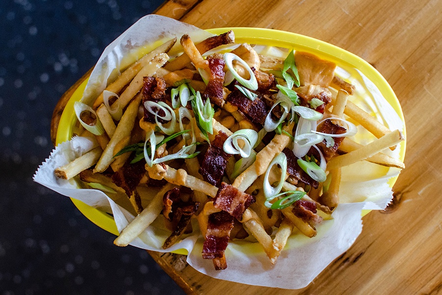 Overhead view of fries topped with bacon, cheese, and scallions and a light wooden table.