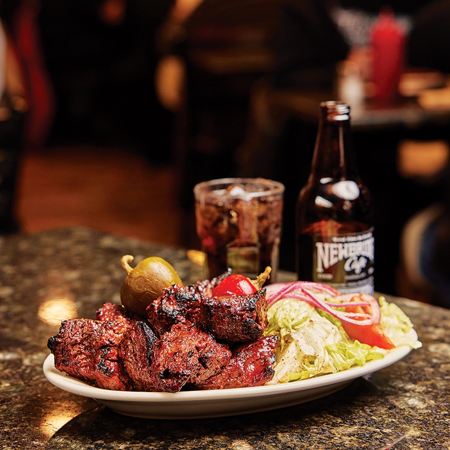 Steak tips, salad, and cherry peppers sit on a plate on a restaurant table, with a glass of soda and its bottle sitting to the site.